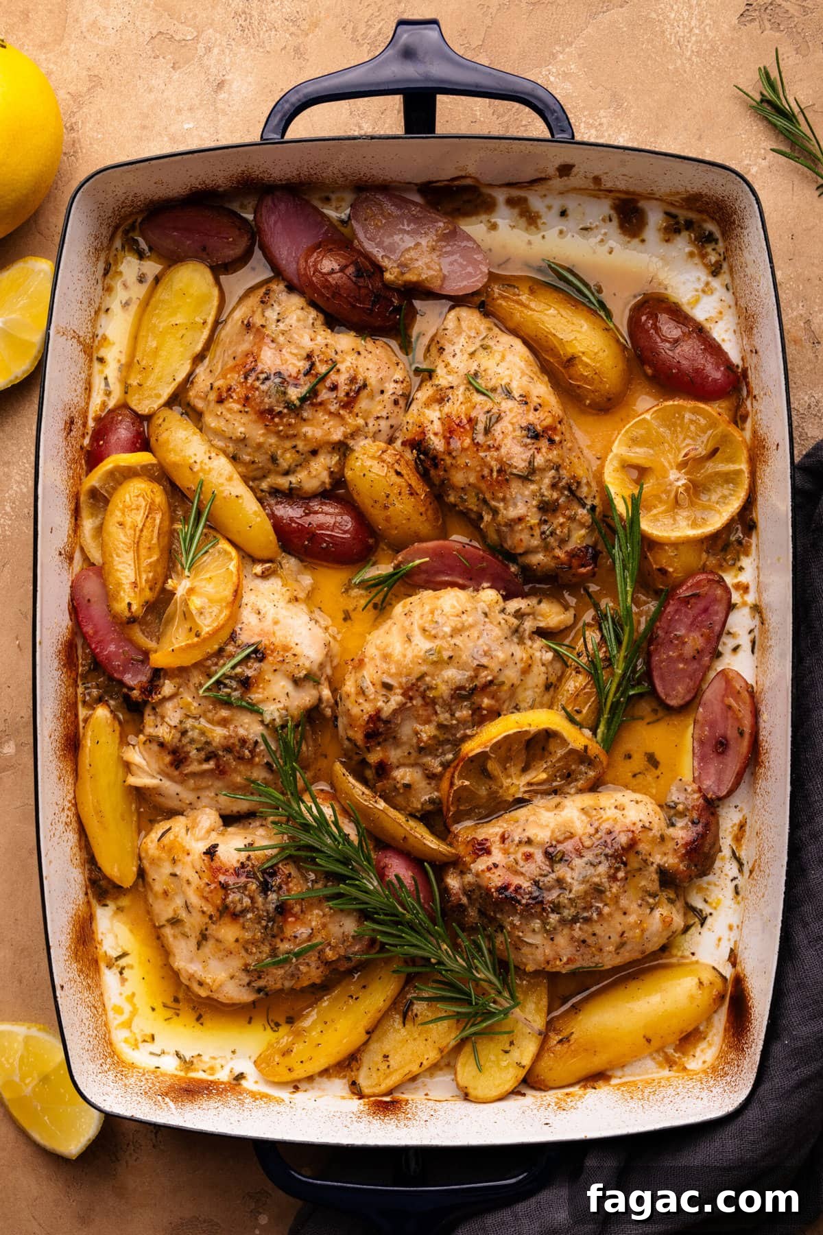 Crispy Roasted Lemon & Rosemary Chicken Thighs with Potatoes 2 Lemon rosemary chicken with potatoes in a rectangle-shaped roasting pan. The pan is displayed on a tan background with lemon slices and a dark gray napkin arranged around it.