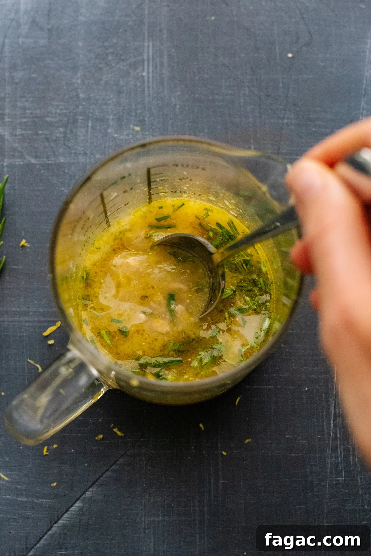 Crispy Roasted Lemon & Rosemary Chicken Thighs with Potatoes 4 hand using spoon to stir lemon sauce in glass measuring cup on gray background.