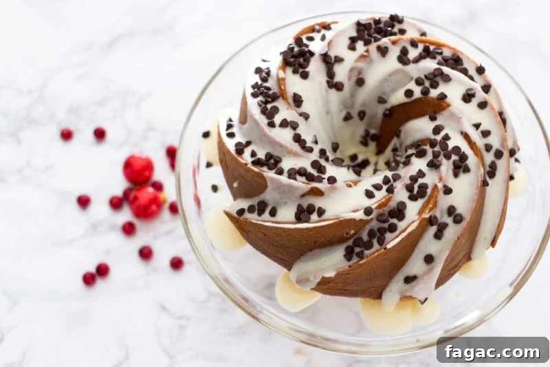 Glass cake dish containing an Orange Vanilla Bundt Cake topped with cream cheese frosting and mini chocolate chips sitting on a marble table.