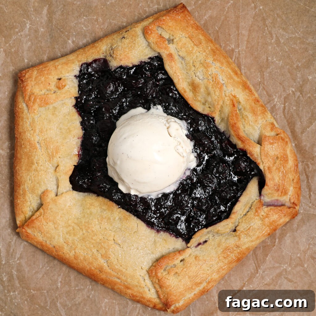 Rustic Blueberry Galette with Buttery Flaky Crust 6 A rustic Blueberry Galette with golden, folded edges, topped with a scoop of vanilla ice cream, sits on a sheet of brown parchment paper.