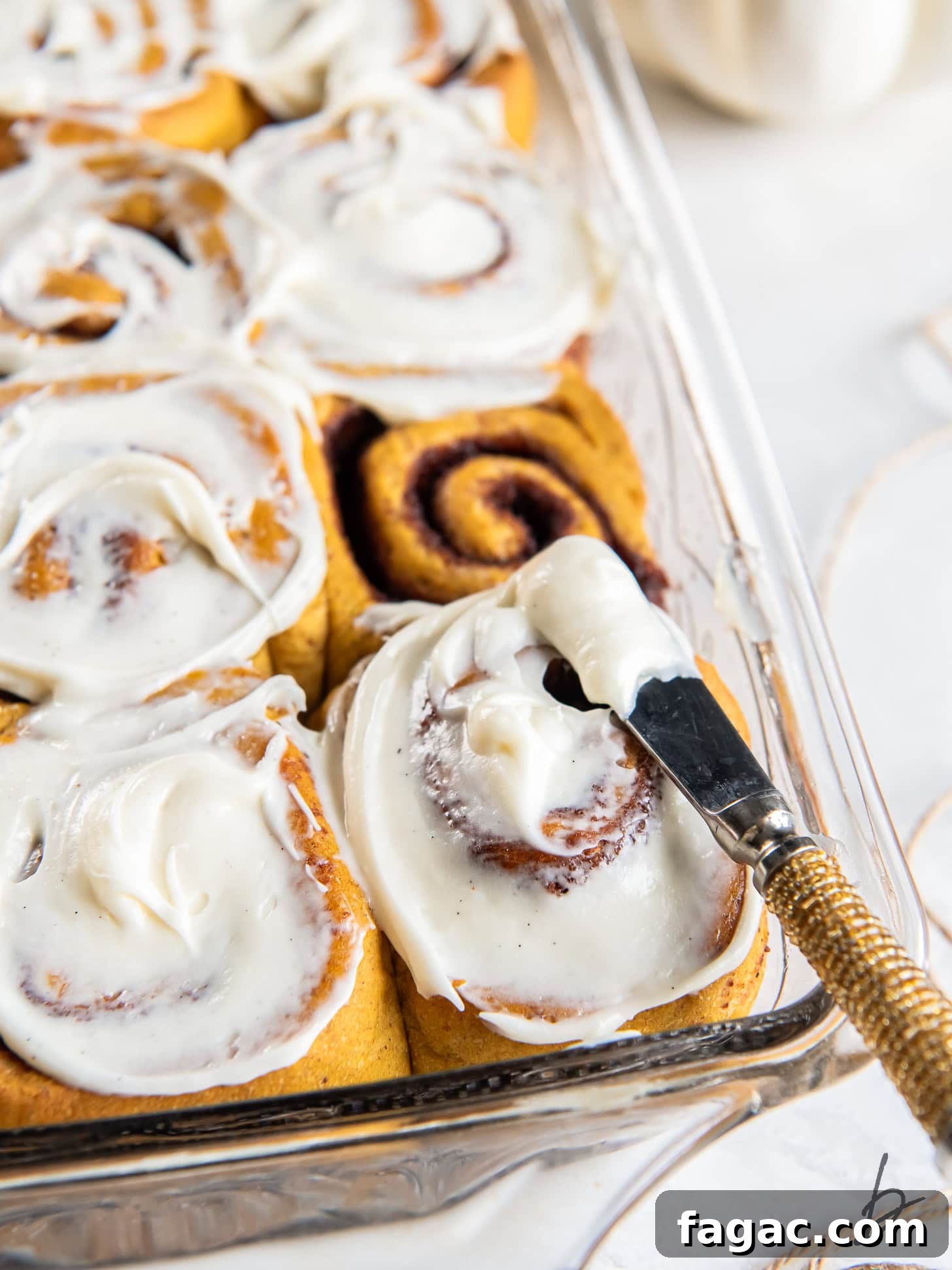 Pumpkin Spice Cinnamon Rolls with Maple Glaze 6 offset spatula spreading cream cheese icing on pumpkin cinnamon rolls in glass baking dish.